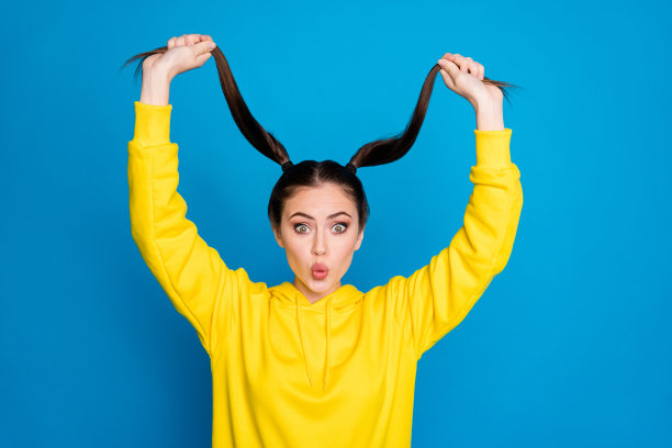 滑稽的漂亮女士的照片举起两个长长的尾巴手臂好心情惊讶快速生长头发穿休闲的黄色连帽衫套头衫孤立亮蓝色背景图片下载