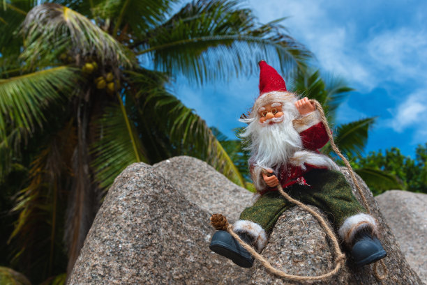 圣诞老人玩具坐在花岗岩与蓝天和棕榈树的背景。完美的新年假期假日概念。图片下载