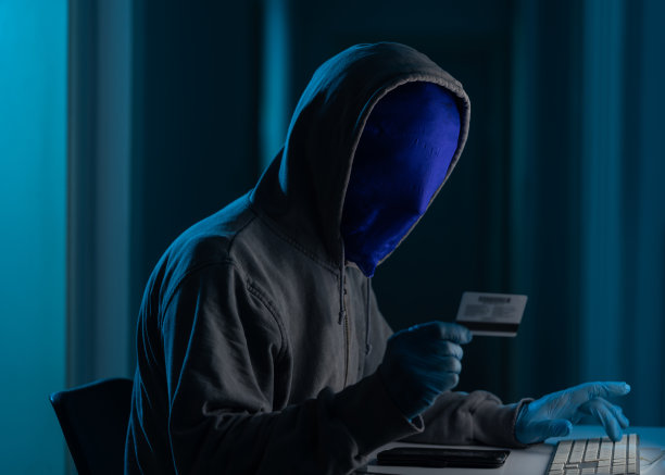 戴着连帽衫和面具的男子在黑暗中使用笔记本电脑进行黑客攻击图片下载