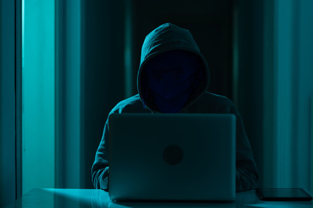 戴着连帽衫和面具的男子在黑暗中使用笔记本电脑进行黑客攻击图片下载