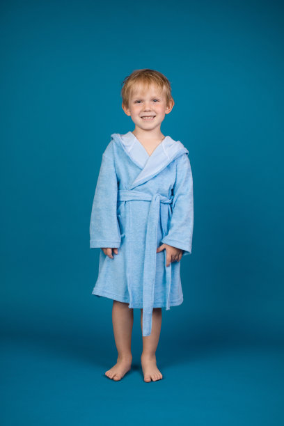 一个迷人的红头发的4岁小孩，穿着淡蓝色的浴袍，带着兜帽和领带，站在那里看着镜头。蒸汽浴、桑拿浴和带游泳池的水疗中心为深蓝色背景。图片下载