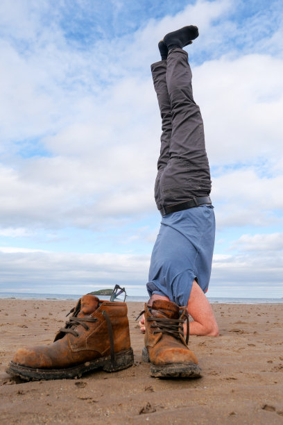 一个男人在海滩上做瑜伽倒立，他的靴子放在头的旁边图片下载