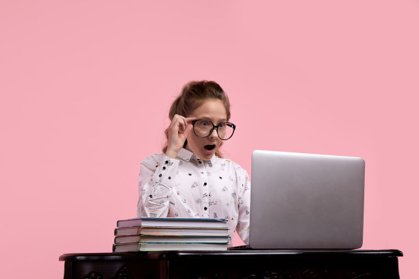惊讶的女孩坐在桌子旁，手里拿着笔记本电脑，一只手拿着眼镜图片下载