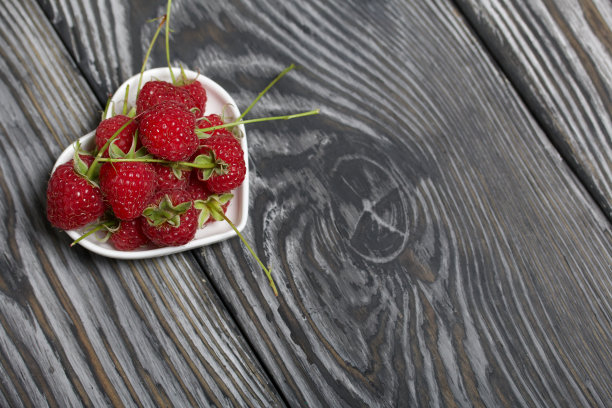 带尾巴的覆盆子躺在一个心形的碟子里。在黑色的黑板上，具有富有表现力的木质纹理。图片下载