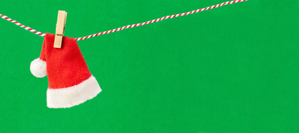 圣诞老人红白相间的皮帽上挂着一根绳子，上面绑着一个木制的晒衣夹。传统象征新年的服装上以绿色横幅为背景，复制空间。图片下载
