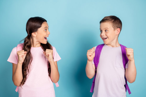 照片欣喜若狂，兴奋的两个小学校的孩子，女孩，男孩印象深刻，收到a级考试成绩，举起拳头，尖叫，是，互相看，穿背包，包，t恤孤立的蓝色背景图片下载