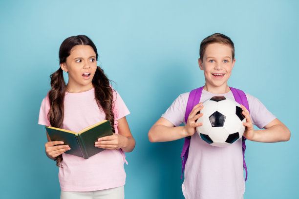 嘿，我不能准备家庭作业。两个小的人的照片生气的孩子女孩极客呆子读者拿书尖叫的男孩踢足球穿休闲t恤包背包孤立的蓝色背景图片下载