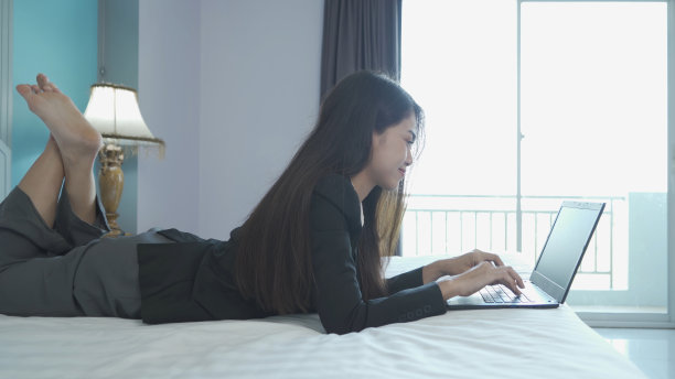 微笑的亚洲泰国商务妇女从家里工作在卧室的床上用电脑笔记本电脑，在电话会议中会见同事在隔离冠状病毒大流行的概念。图片下载