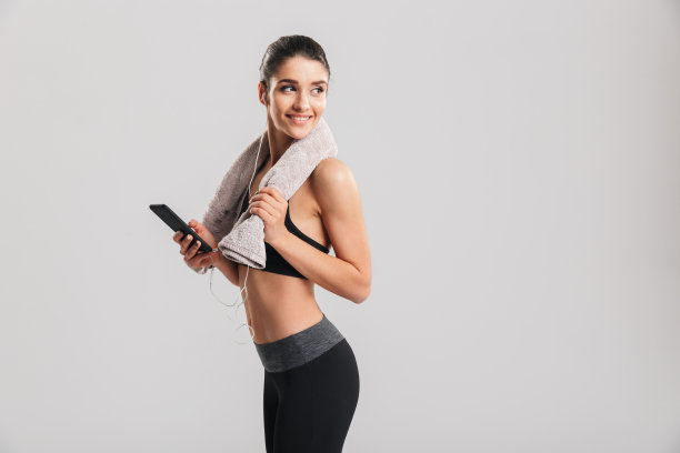 照片上的女人微笑着穿着运动服，通过耳机听音乐训练后，脖子上的毛巾，在灰色的背景下孤立图片下载