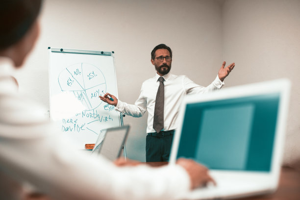 局长正在向同事们提交一份商业计划。一个打着领带穿白衬衫的人拿着一张图表站在黑板前。办公室职员在开会时坐在一张桌子前，桌上放着笔记本电脑。高质量的照片图片下载