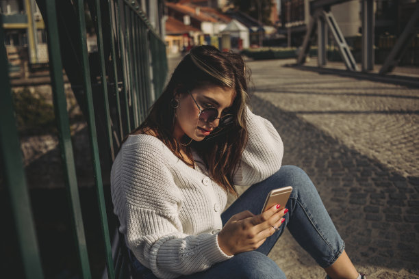 一个时髦的年轻女孩正坐在城市的一座桥上用手机休息图片下载