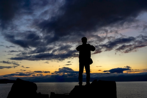 一个在山顶看日落的无名男子，日出或日落时在外面拍照的男子剪影图片下载
