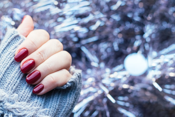 女性手在灰色针织毛衣与美丽光滑的指甲-酒红色，深红色，樱桃色的指甲在背景的淡粉色圣诞金丝花环与复制空间。有选择性的重点图片下载