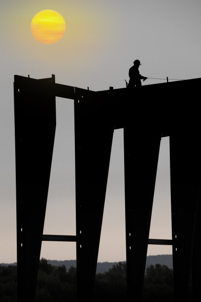在冉冉升起的太阳下，一个工人正在建造一座钢铁工业建筑。图片下载