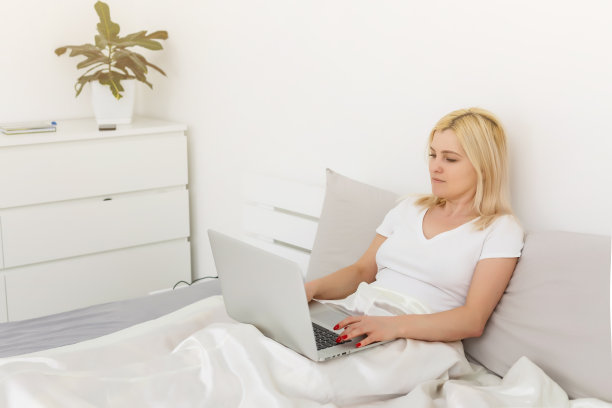 在酒店房间工作。美丽的年轻女商人工作在笔记本电脑和微笑，而坐在床上的酒店房间图片下载