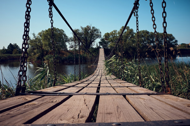 河上有一座悬索桥，通往岛上，有落叶树。铁链之间系着木条。扶手由金属电缆。绕着绿色的芦苇。图片下载