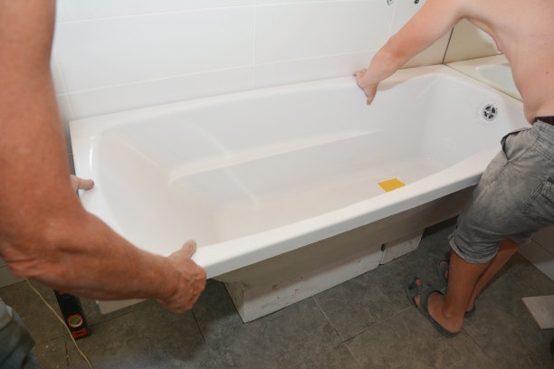 建筑承包商正在一间新铺瓷砖的浴室的角落里，吊装一个白色丙烯酸标准浴缸。图片下载