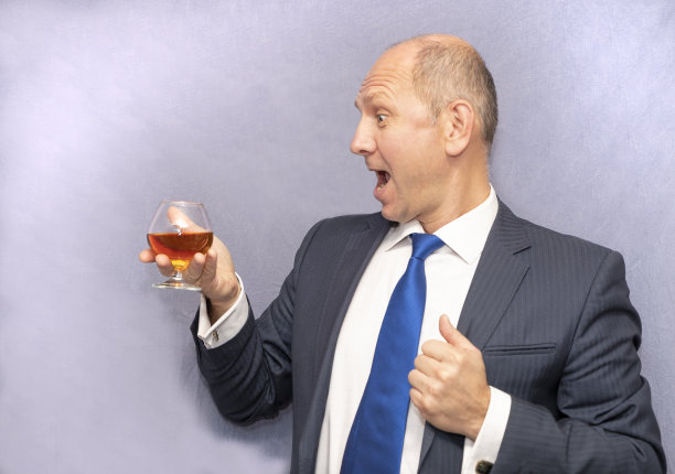 脸上洋溢着喜悦的情绪。一个高加索人，喝着一杯白兰地。一个穿着西装，背景是灰色的男人。图片下载