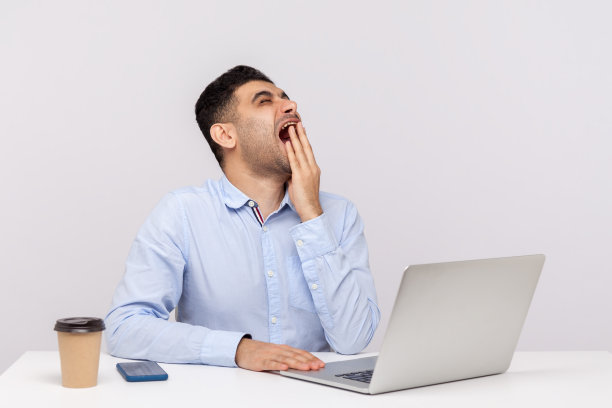 疲惫的男人在办公室工位用笔记本电脑工作，打哈欠并用手捂住脸图片下载