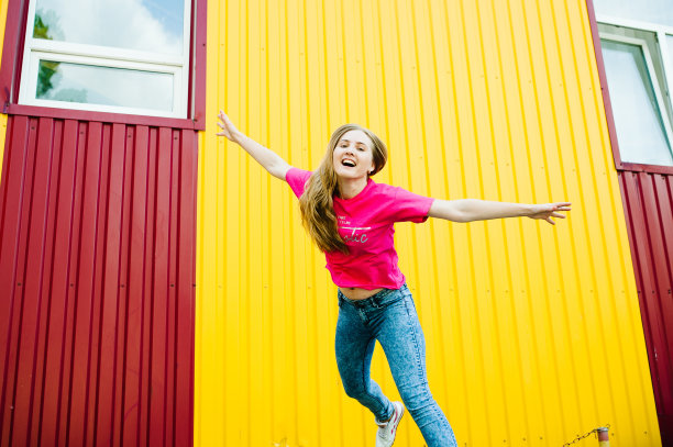 美丽的运动的年轻女子，长金发，粉红色的衬衫和蓝色的牛仔裤。在黄红色背景的车库墙摆姿势和微笑。使跳跃,飞。风把头发图片下载