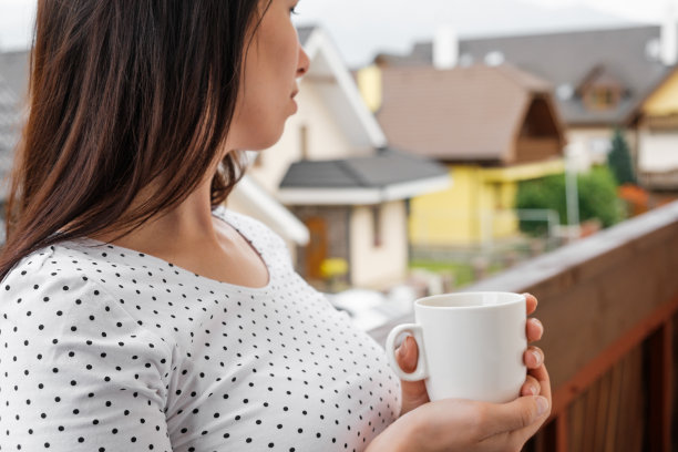 年轻女子穿着圆点t恤，端着一杯白色的茶或咖啡站在阳台上。乡村的早晨。放松的概念。图片下载