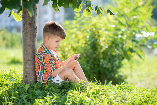 一个美丽的孩子坐在草地上看手机在夏天日落。手里拿着手机的可爱男孩图片下载