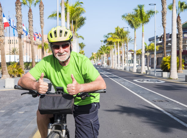 一位戴着眼镜和黄色头盔的老人在加那利群岛特内里费荒凉的道路上骑着自行车。2019冠状病毒病导致游客减少。背景是棕榈树和蓝天图片下载