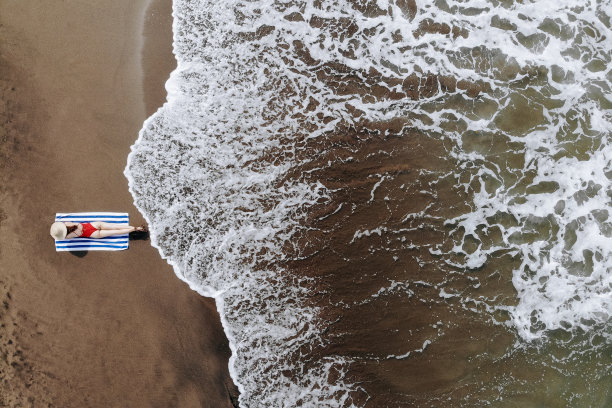 顶视图的女人在红色比基尼和草帽上晒黑的毛巾在海滩上的黑色的沙子。在巴厘岛度假。从无人机的照片图片下载