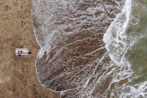 鸟瞰图上的妇女日光浴红色比基尼和草帽上晒黑的毛巾在海滩上与黑色的沙子。在巴厘岛度假。从无人机的照片图片下载