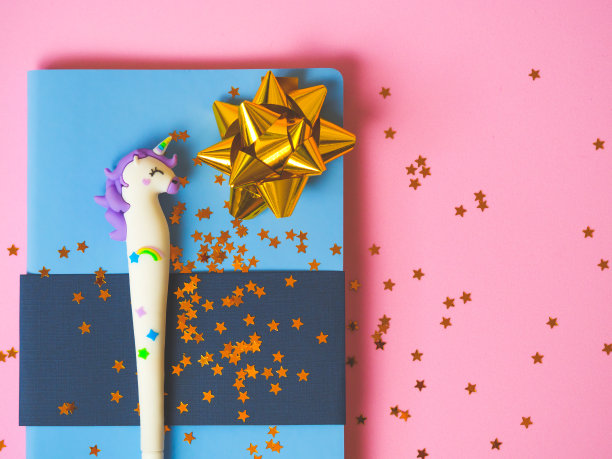蓝色笔记本，可爱的独角兽笔，金色的蝴蝶结，粉红色的背景上闪烁着星星。图片下载