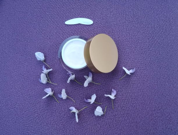 白色的化妆品(面霜或身体霜)在开放的透明闪亮的罐子，金色的盖子，白色的涂抹器和紫藤花瓣在紫罗兰纹理的背景图片下载