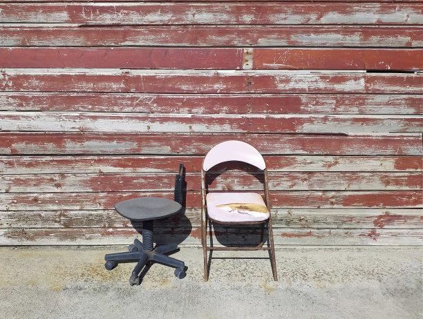 两张生锈坏了的旧椅子:一张粉红色的可折叠椅子和一张有轮子的桌子，靠在一堵没有塞上软木塞的木板墙上。物质事物的原始成分的变质。图片下载