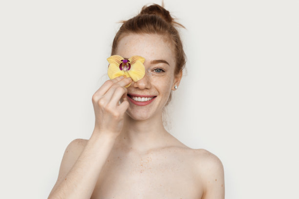 微笑的白人女士与红头发和雀斑覆盖的眼睛与一朵花和摆姿势的赤裸肩膀在一个工作室的白色墙壁图片下载