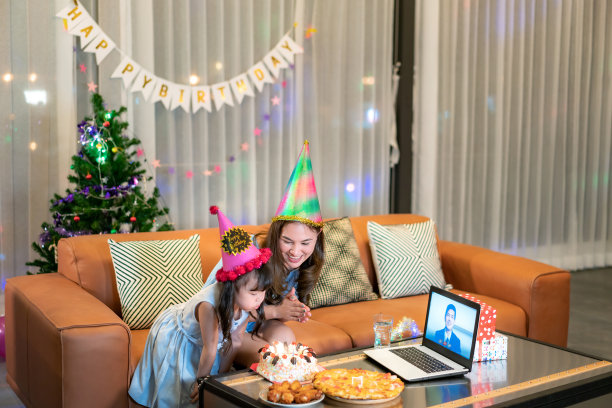 快乐的小女孩和妈妈在家庆祝生日，爸爸通过笔记本电脑视频通话，吹灭桌上的蛋糕蜡烛图片下载