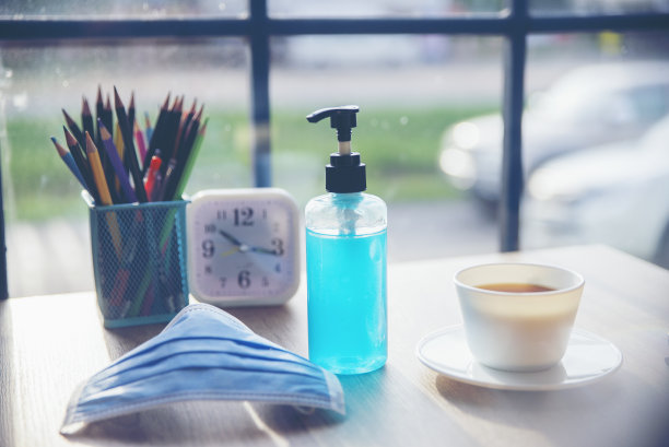 新常态与防冠状病毒(洗手液)概念。酒精凝胶和卫生口罩用于隔离covid-19。咖啡杯，钟表和铅笔放在家庭办公室的木桌上。图片下载