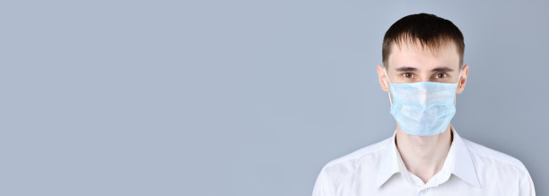 横幅。一个戴医用口罩的男人的特写。一个英俊的黑头发，棕色眼睛的男人，穿着白色衬衫和医用口罩，睡袍看着相机，在一个灰色的背景。在左边有一个空白区域用于文本。冠状病毒，Covid-图片下载