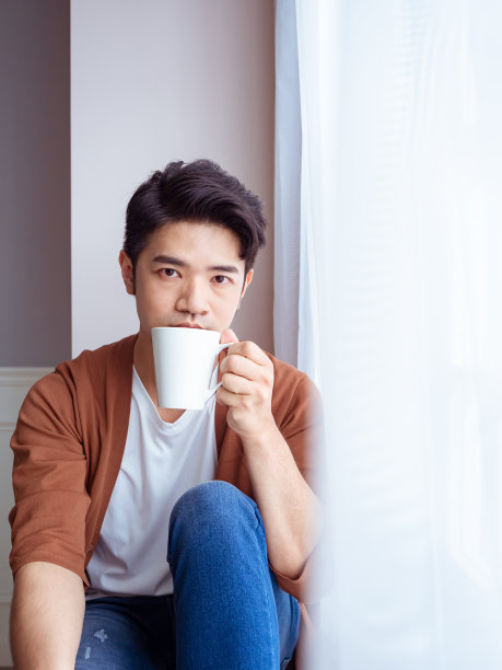 在窗帘旁饮酒的英俊年轻男人图片下载