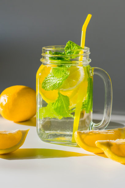 一张特写的垂直照片，一个杯子装满水，切片柠檬和薄荷叶与黄色吸管，旁边是一片柠檬图片下载
