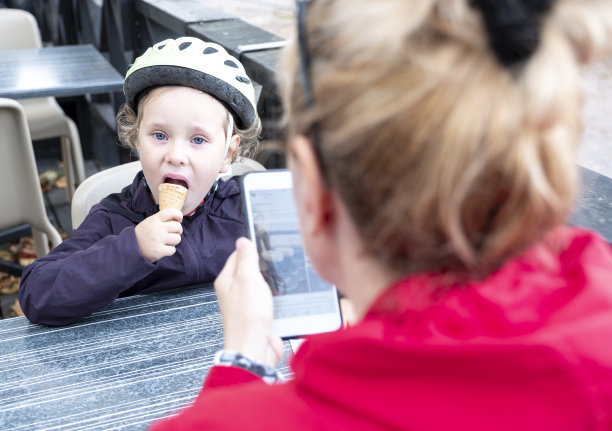外面咖啡馆里的女人和女孩。小女孩吃着冰淇淋，妈妈用智能手机拍下了她的照片。图片下载