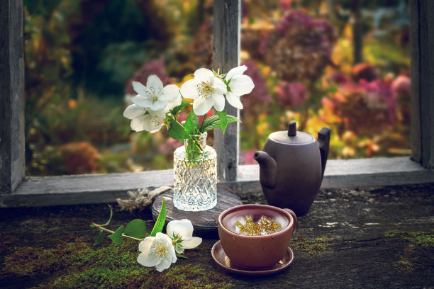 旧窗边的旧木桌上放着茉莉花茶，窗外是秋天的风景。茶杯里的茶，茶壶和花瓶里的花图片下载