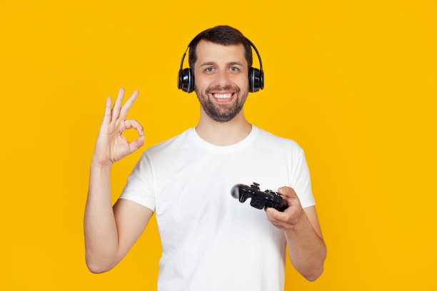 一个穿着白色t恤留着胡子的年轻人，戴着耳机玩电子游戏，用手指做一个正常的手势，这是一个很好的象征。站在孤立的黄色背景上图片下载