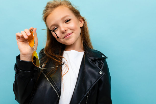 迷人的欧洲女孩在皮夹克拿太阳镜在她的手在浅蓝色的背景图片下载