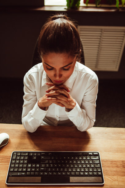 一位女职员坐在她的办公桌前，双手合十。女人看着桌子上的电脑键盘。高质量的照片图片下载