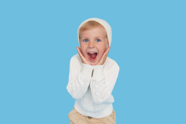 哇的脸。可爱的金发小男孩，戴着温暖的白色帽子，穿着蓝色背景的毛衣。冬季儿童打折销售图片下载