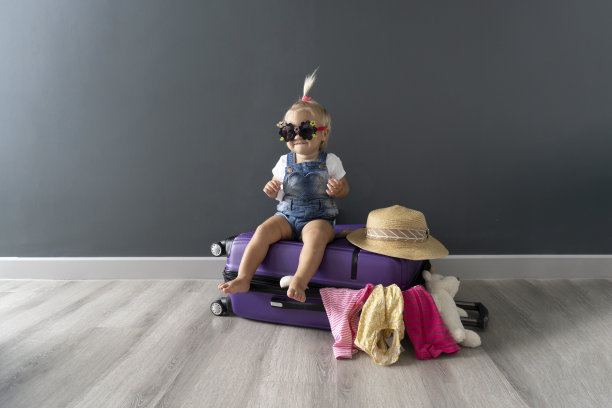 光着脚、戴着黑色太阳镜的可爱金发小女孩坐在一个紫罗兰色的行李箱上，等待着暑假去国外旅游。图片下载