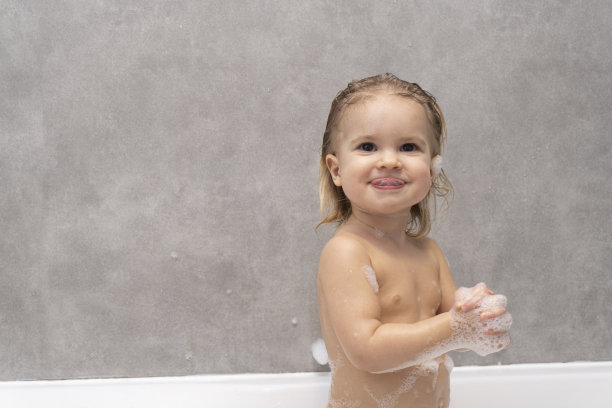 可爱的小白种婴儿微笑着在浴室里洗着泡沫浴，灰色的背景，复制空间，快乐的童年概念。图片下载