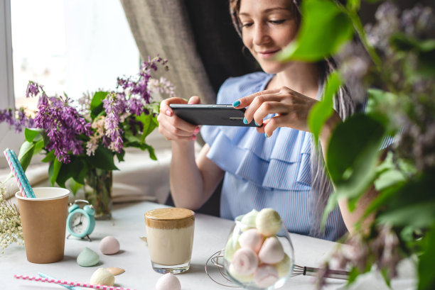 一名妇女正在用手机拍下一杯清晨的达尔戈纳咖啡。白色背景，柔和的颜色和淡紫色的花图片下载