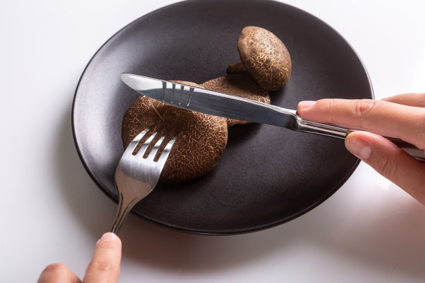 一个人拿着刀叉刀叉，在白色背景的黑色盘子上切着生的泽西牛蘑菇。手在画框里。水平方向。高质量的照片图片下载