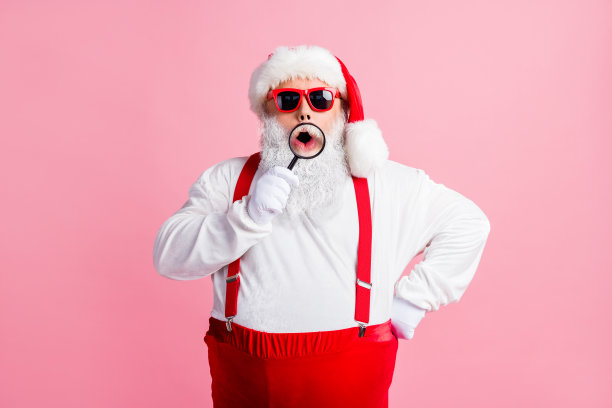 照片胖惊讶的圣诞老人拿着放大镜搜索传统的圣诞节折扣印象深刻的张开嘴戴时髦的太阳镜吊带帽孤立的彩色背景图片下载