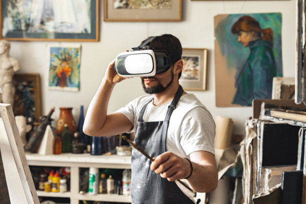 男画家在艺术工作室室内戴着虚拟现实耳机拿着画笔查看图片，好奇图片下载